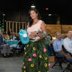 Habitat Greensboro board member, Amina, smiles wearing a long skirt of branches and flowers. She looks off screen while holding a blue watering can.