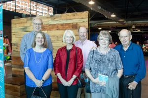 A group of Habitat Greensboro stakeholders stand smiling together in front of a wooden background at Blueprints and Bubbly.