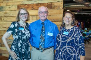 One man dressed in a blue shirt smiles between two women wearing blue dresses.