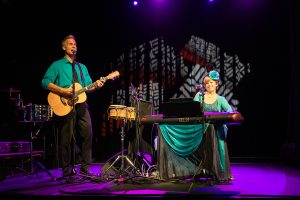 Two band members perform on stage wearing blue attire. The man sings while holding a guitar while the woman smiles and plays a keyboard.