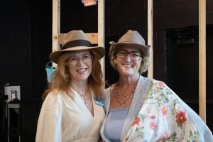 Two women wearing glasses and tan fedoras smile and pose next to each other while standing in a half embrace.