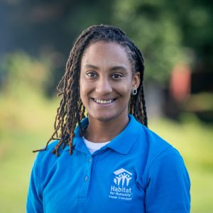 A woman with shoulder-length, curly, brown locs smiles at the camera while wearing a Habitat Greensboro blue polo t-shirt.