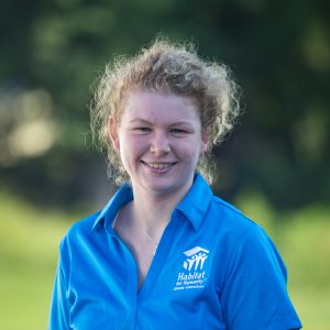 A woman with curly, blond hair in a bun and blue-eyes smiles at the camera while wearing a Habitat Greensboro blue polo t-shirt.