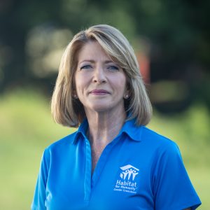 A woman with shoulder-length blond hair and blue-eyes smiles at the camera while wearing a Habitat Greensboro blue polo t-shirt.