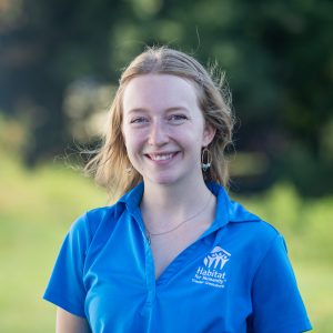 A woman with blond, straight hair smiles at the camera while wearing a Habitat Greensboro blue polo t-shirt.