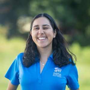 A woman with dark brown, wavy hair smiles at the camera while wearing a Habitat Greensboro blue polo t-shirt.