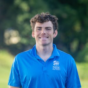 A man with short, curly, brown hair smiles at the camera while wearing a Habitat Greensboro blue polo t-shirt.