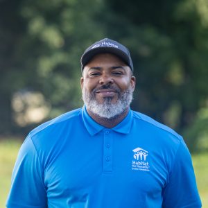 A man with a curly, grey beard smiles at the camera while wearing a Habitat Greensboro grey ball cap and blue polo t-shirt.