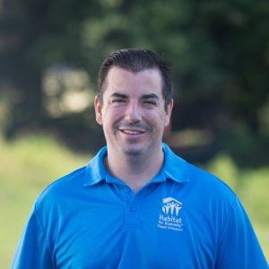 A man with short, black hair and dark eyebrows smiles at the camera while wearing a Habitat Greensboro blue polo t-shirt.