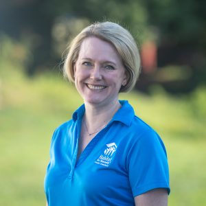 A woman with ear-length, straight, blonde hair smiles at the camera while wearing a Habitat Greensboro blue polo t-shirt.