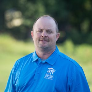 A man with buzzed, brown hair smiles at the camera while wearing a Habitat Greensboro blue polo t-shirt.