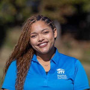 A woman with brown, braided hair smiles at the camera while wearing a Habitat Greensboro blue polo t-shirt.