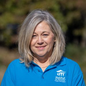 A woman with grey hair smiles at the camera while wearing a Habitat Greensboro blue polo t-shirt.