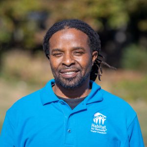 A man with long, black locs smiles at the camera wearing a Habitat Greensboro blue polo shirt.