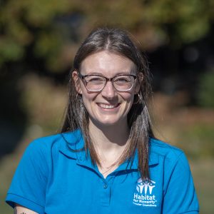 A woman smiles at the camera wearing glasses and a blue polo shirt.